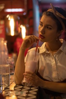 A woman with red lips and headband drinking a milkshake in a dimly lit cafe.