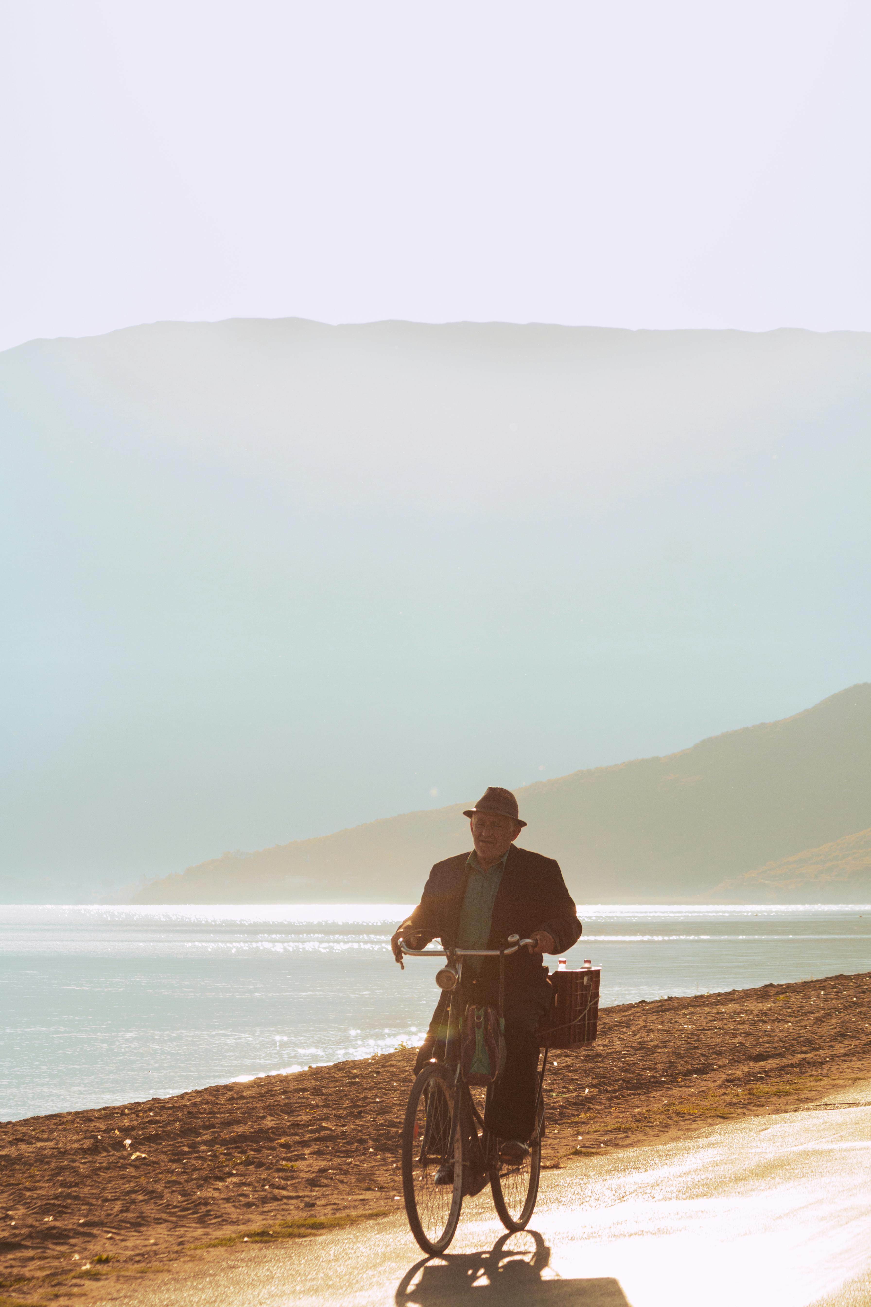 Elderly man on bicycle rides alongside a peaceful mountain lake under clear skies.