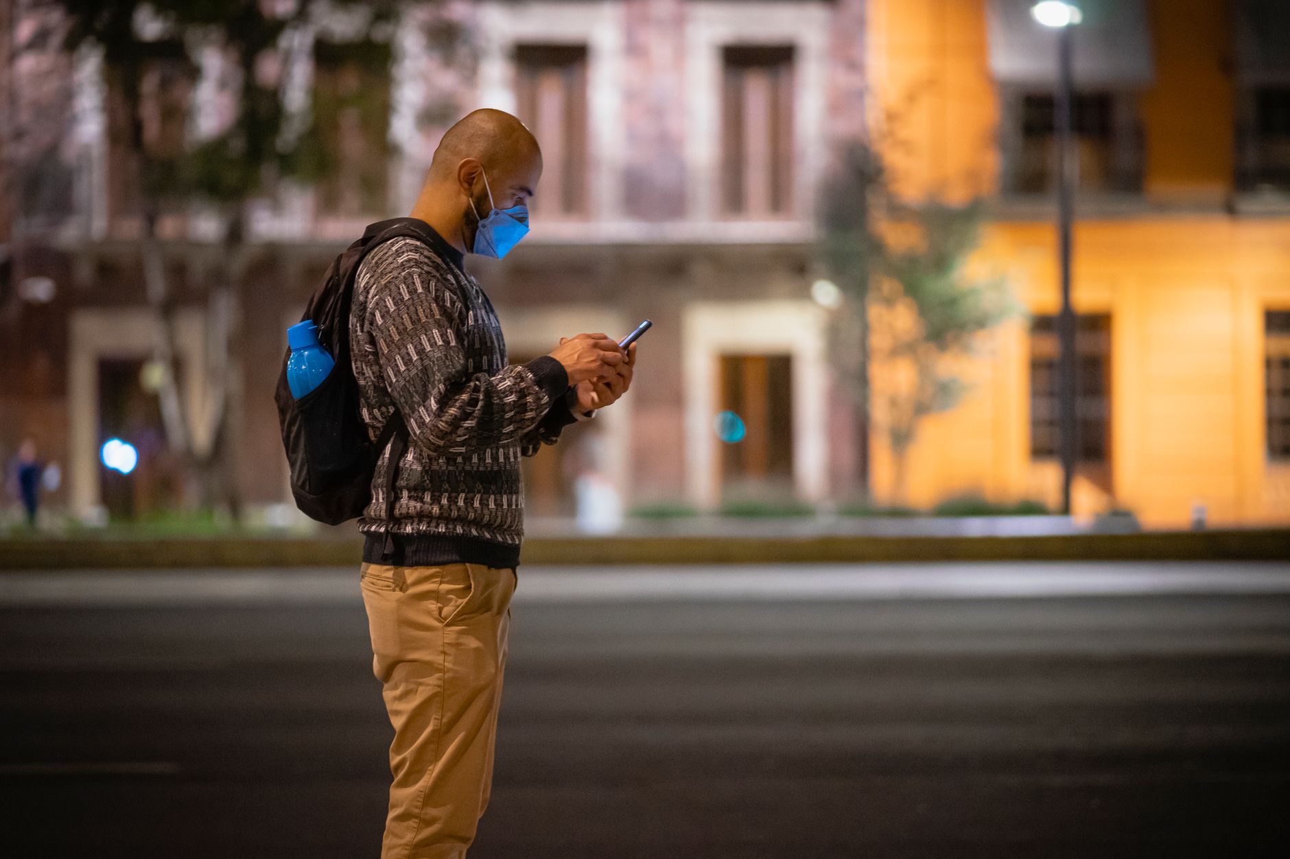 A man using a smartphone at night on a street in Mexico City, wearing a face mask.