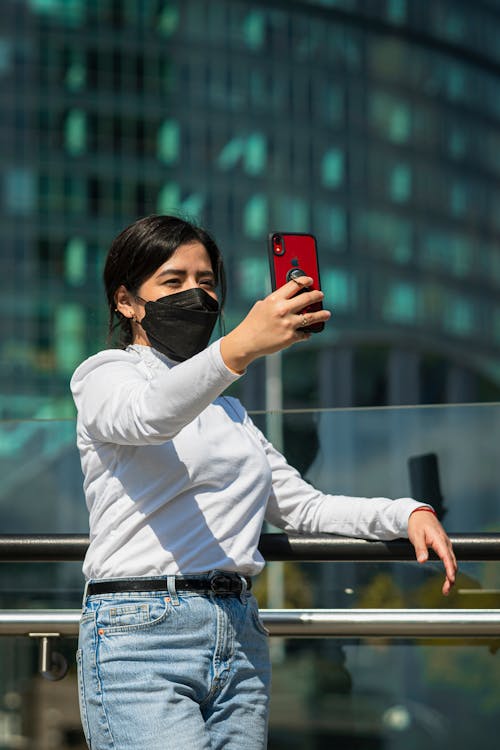 Free Woman in White Long Sleeve Shirt and Blue Denim Jeans Holding Red Smartphone Stock Photo