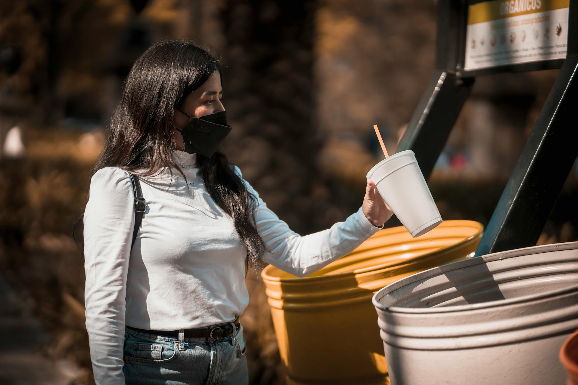Woman in mask disposes paper cup in outdoor recycling bin on a sunny day.
