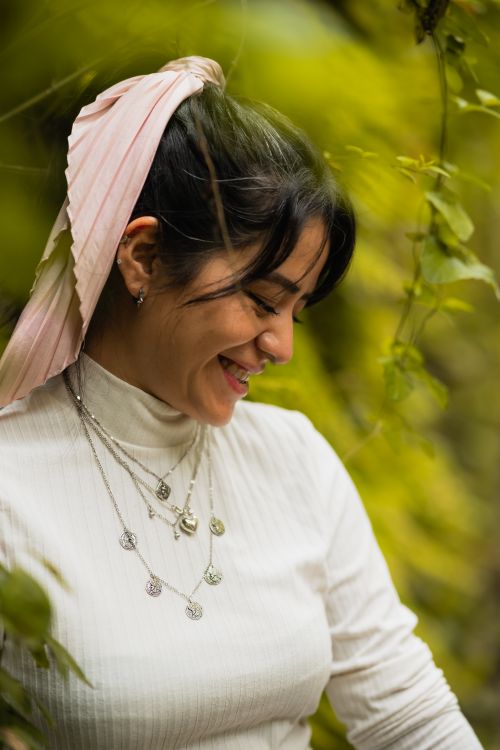 Free A young woman with a pink scarf smiles amidst lush greenery, showcasing layered necklaces outdoors. Stock Photo