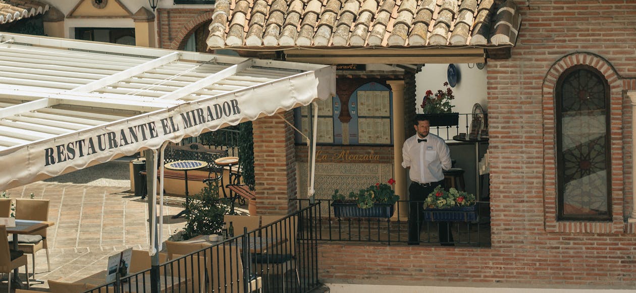 Spanish restaurant with terrace awning labeled "Restaurante El Mirador" and waiter standing at brick entrance.