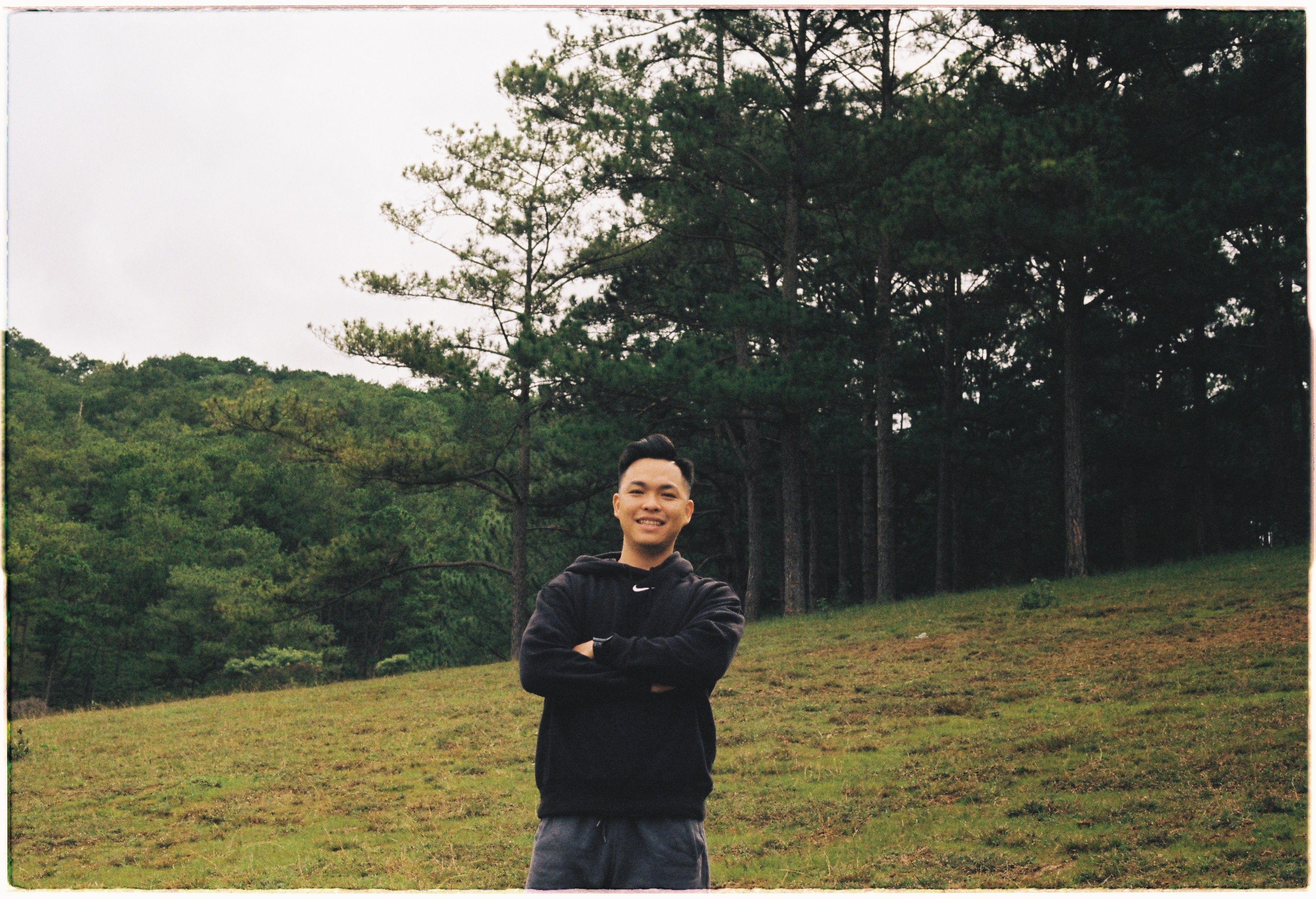 Young Asian man smiling with arms crossed, standing in a serene forest setting.