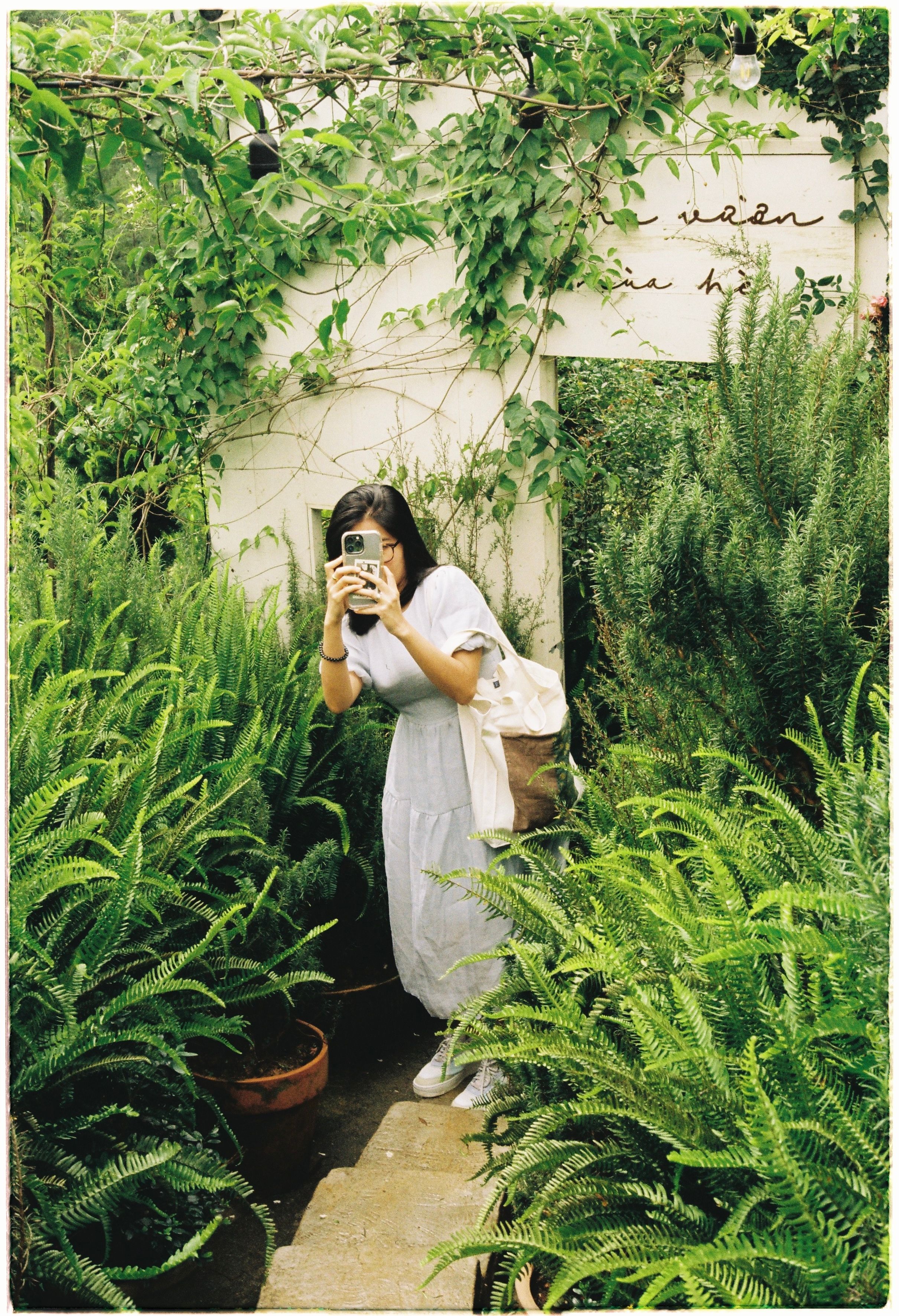 A woman with black hair takes a photo using her phone in a lush, vibrant garden setting.