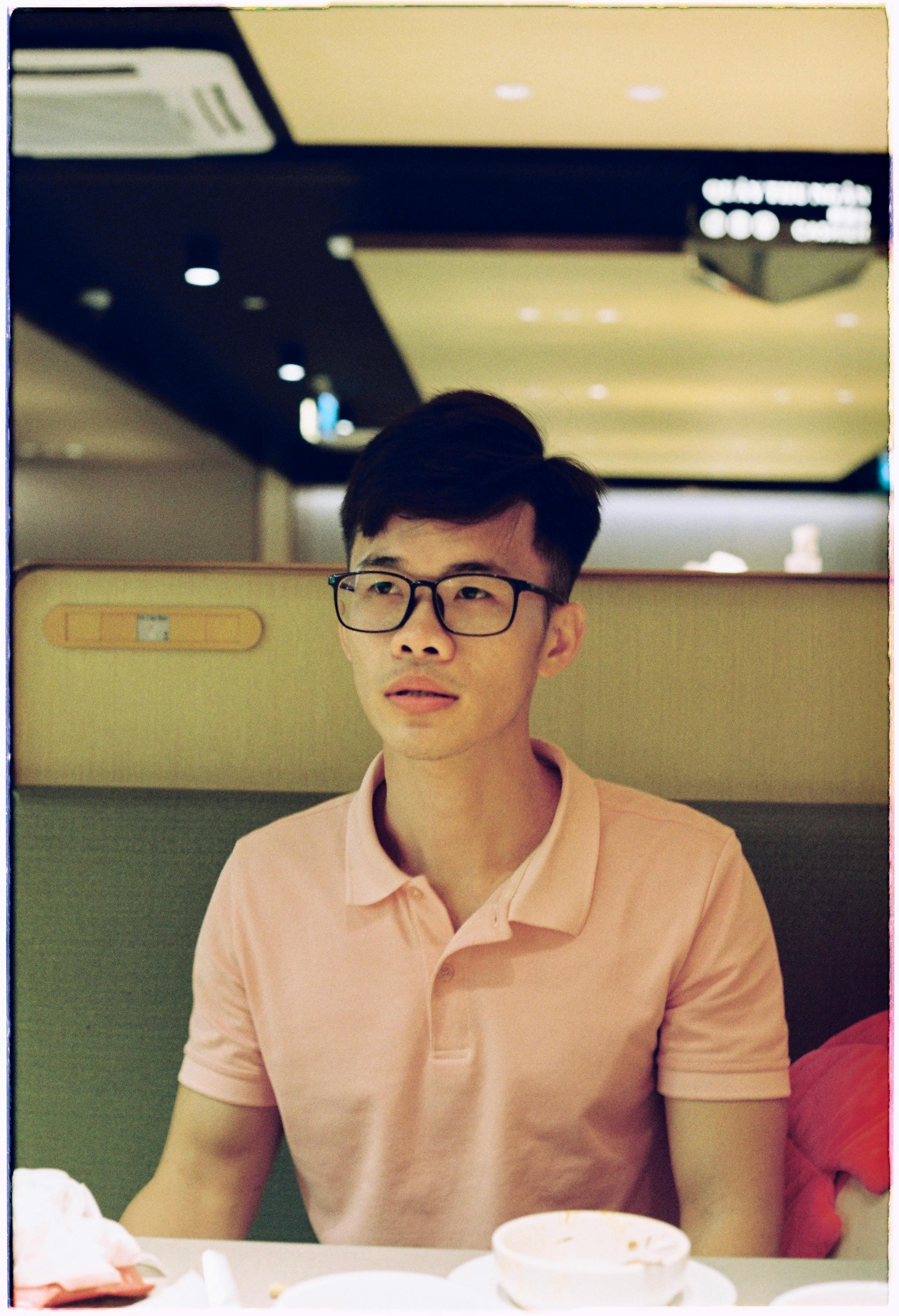 Portrait of a man wearing glasses and a pink shirt, sitting in an indoor dining setting.