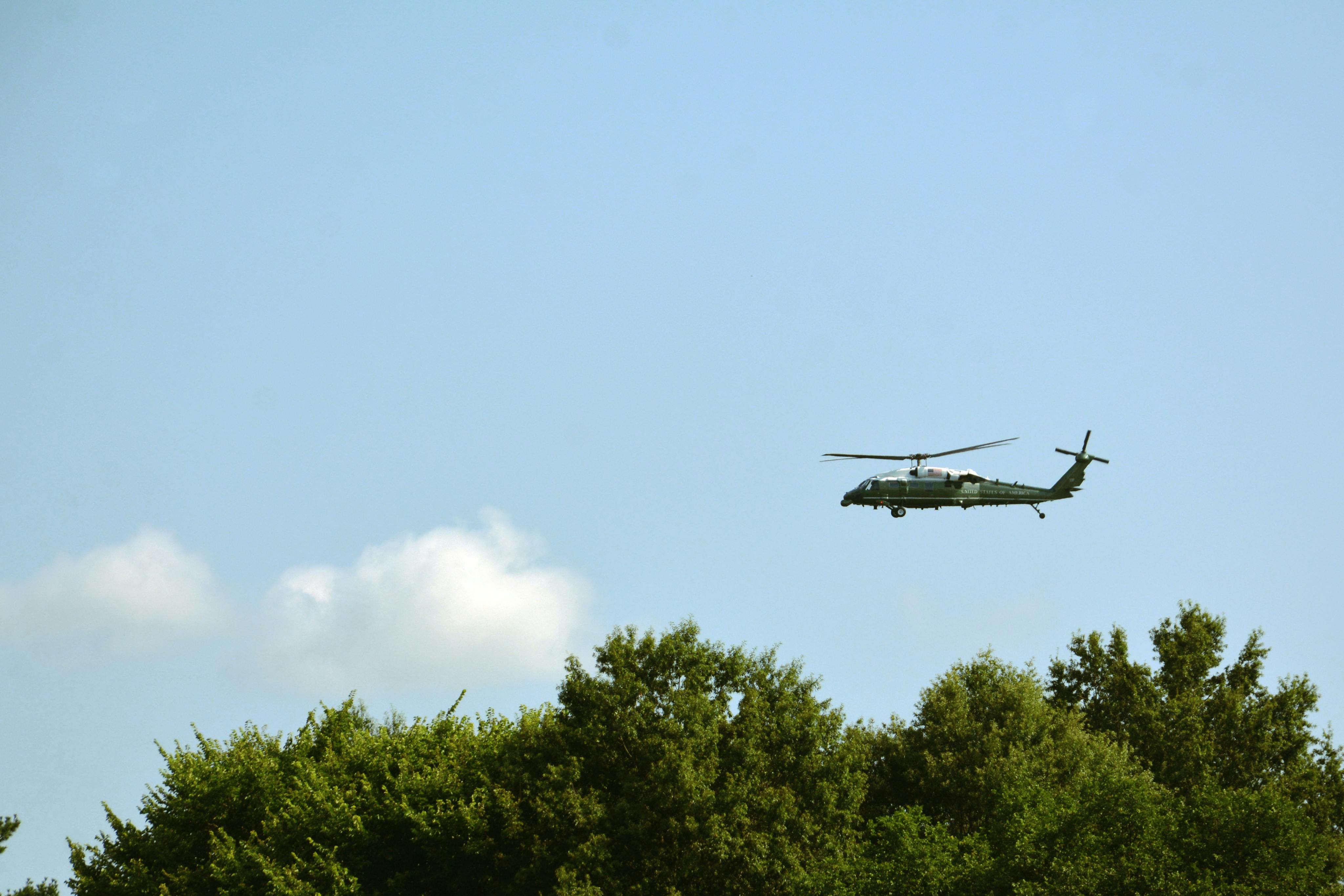 Utility helicopter flying high above green trees in Washington, DC under a clear sky.