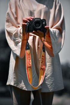 A person in a light-colored shirt holds a Zenit camera outside, featuring a bold strap.