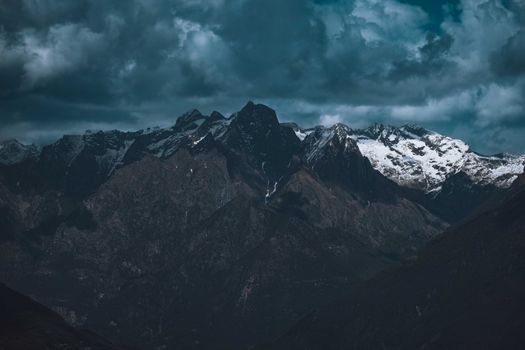 Dramatic view of rugged snow capped mountains beneath a dark, cloudy sky, conveying a moody and atmospheric scene.