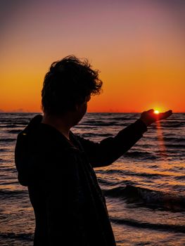 A person holds the setting sun in their hand on Jūrmala beach, Latvia, during a vibrant sunset.