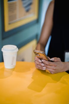 A woman texts on her phone at a cafe, a coffee cup resting on a vibrant yellow table.