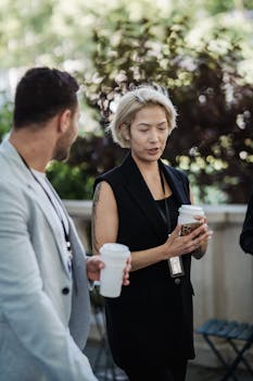 Two adults engaged in conversation outdoors while holding coffee cups, in a professional setting.