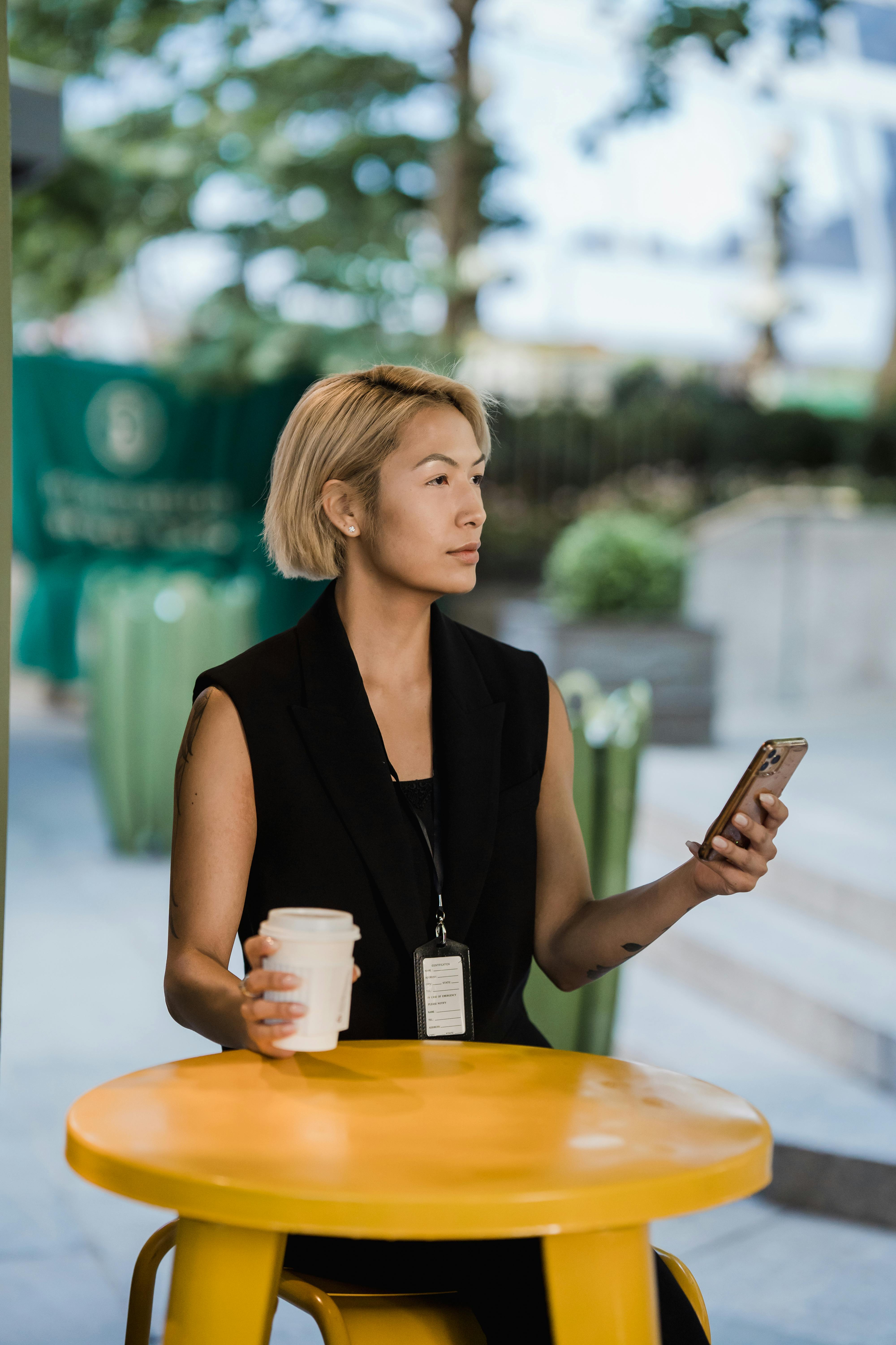 woman with an identification badge sitting behind a table and holding a phone and a coffee cup