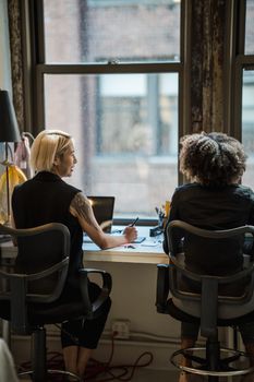 Two women working together at an office desk by a window, discussing ideas in a relaxed setting.