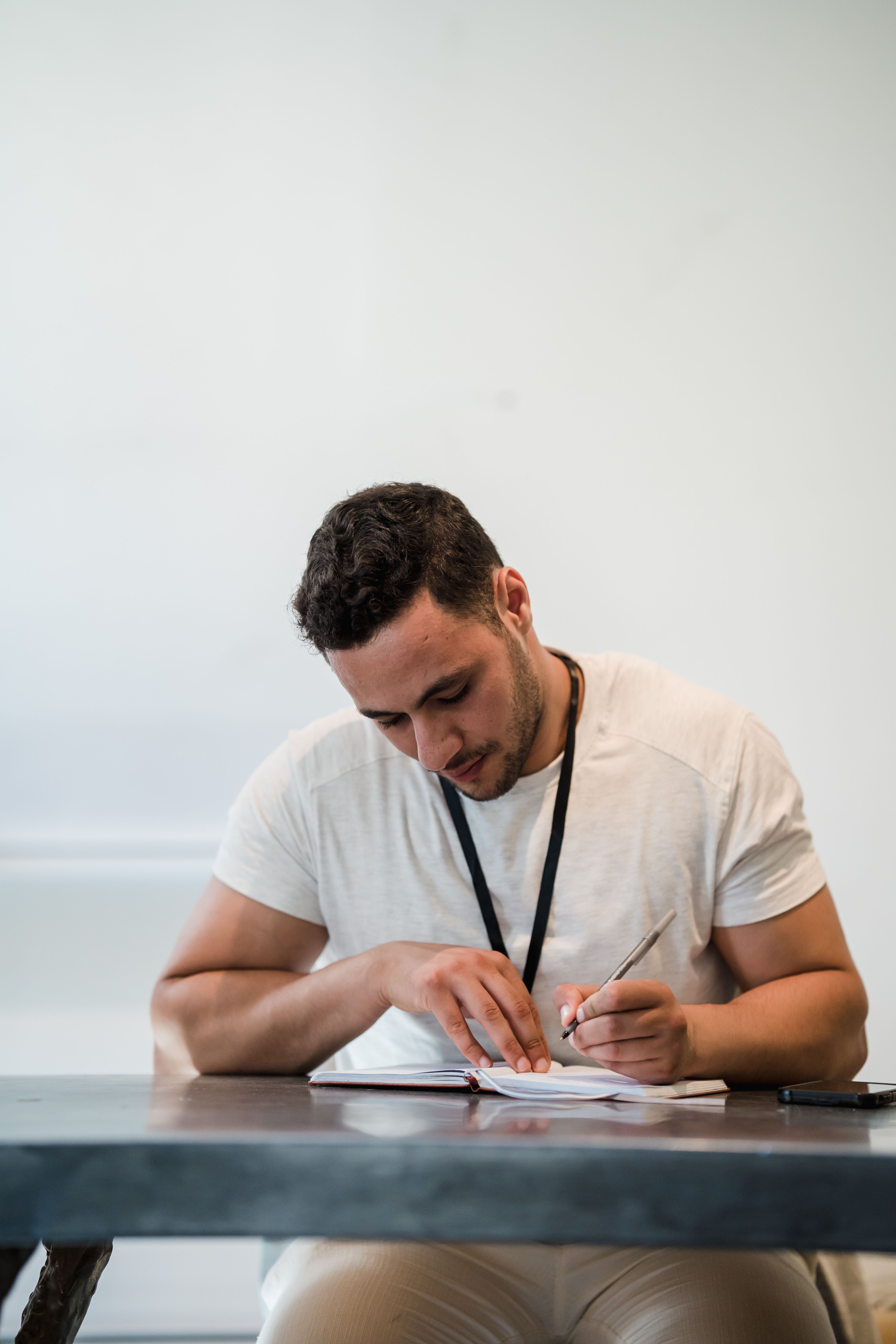 Adult man with stubble writing in a notebook at a desk. Modern and minimalistic setting indoors.