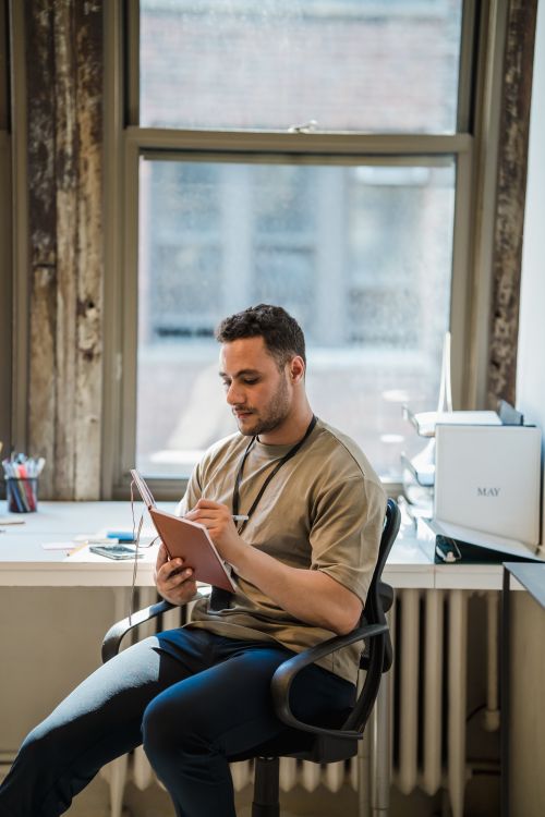 Free A young man sitting at a desk by the window, focused on writing in a notebook in a well-lit office. Stock Photo