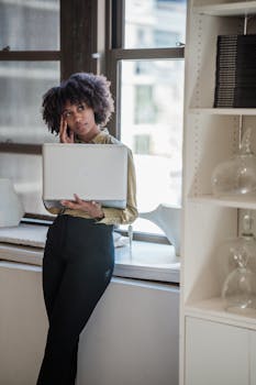 Businesswoman leaning on a windowsill, holding a laptop in a sunlit office.