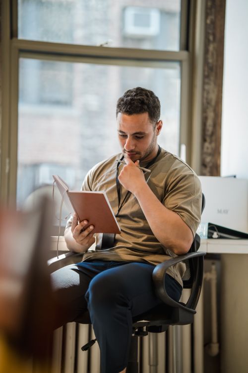 Free Man sitting by a window in an office, writing notes in a notebook. Thoughtful and engaged. Stock Photo