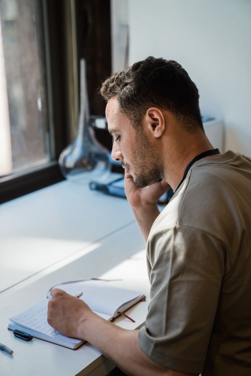 Free Man writing in a notebook by a sunlit window, thoughtful and focused. Stock Photo