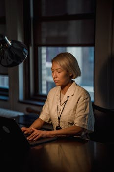 Asian woman working on a laptop in a dimly lit office with a desk lamp for focus.
