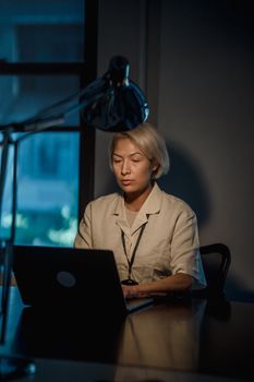 A woman with blonde hair works intently on a laptop in a dimly lit office.