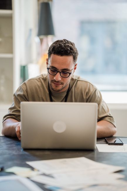 Man in glasses concentrating on work at desk with laptop in bright office setting.