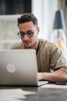 Young man in glasses intensely working on a laptop at a desk indoors.