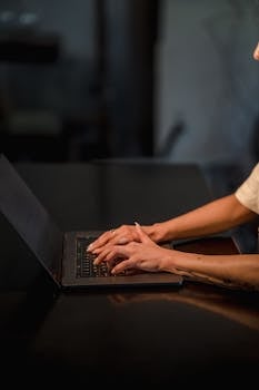 A woman typing on a laptop in a dimly lit setting, focusing on her hands.