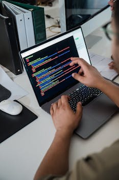 Close-up of a programmer pointing at a colorful code script on a laptop in an office setting.