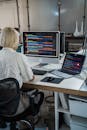 Back View of a Blond Woman in an Office Looking at Two Computer Screens with Multicoloured Code