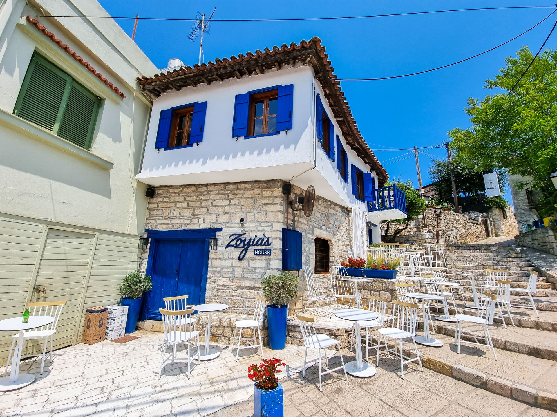 Outdoor dining area at a traditional stone tavern with blue shutters in Afytos, Greece under a clear sky.