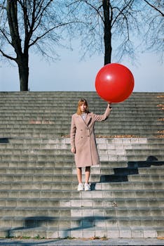 Stylish woman in coat holding a red balloon on urban steps with leafless trees in background.