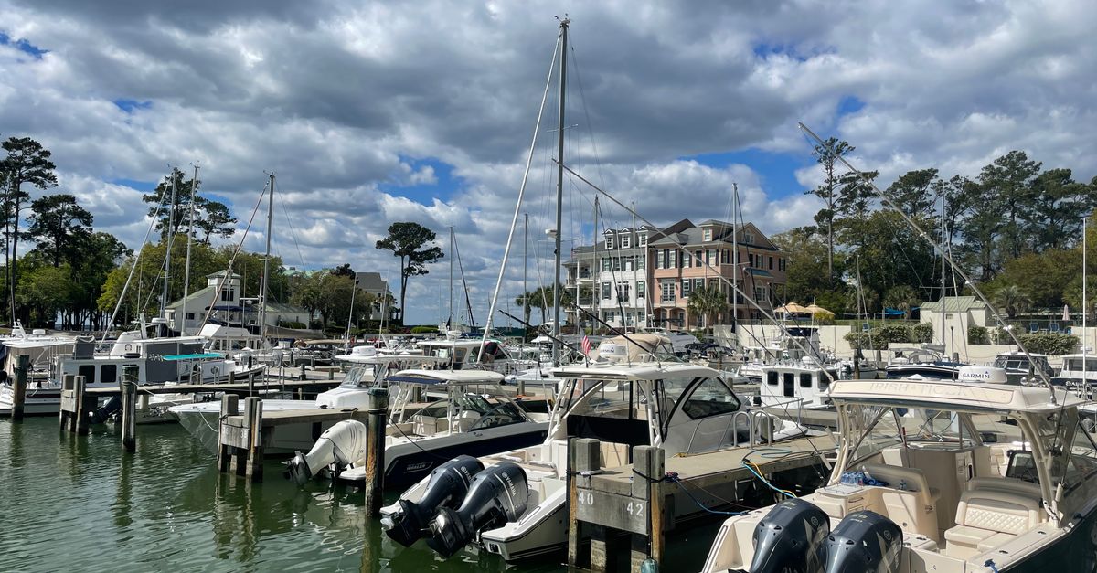 Explore the tranquil marina at Hilton Head Island with docked sailboats and motorboats under a partly cloudy sky.