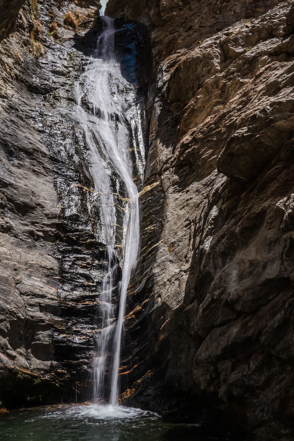 A tall thin waterfall cascading down rugged rock walls into a narrow gorge, similar to Manai Falls at Takachiho Gorge