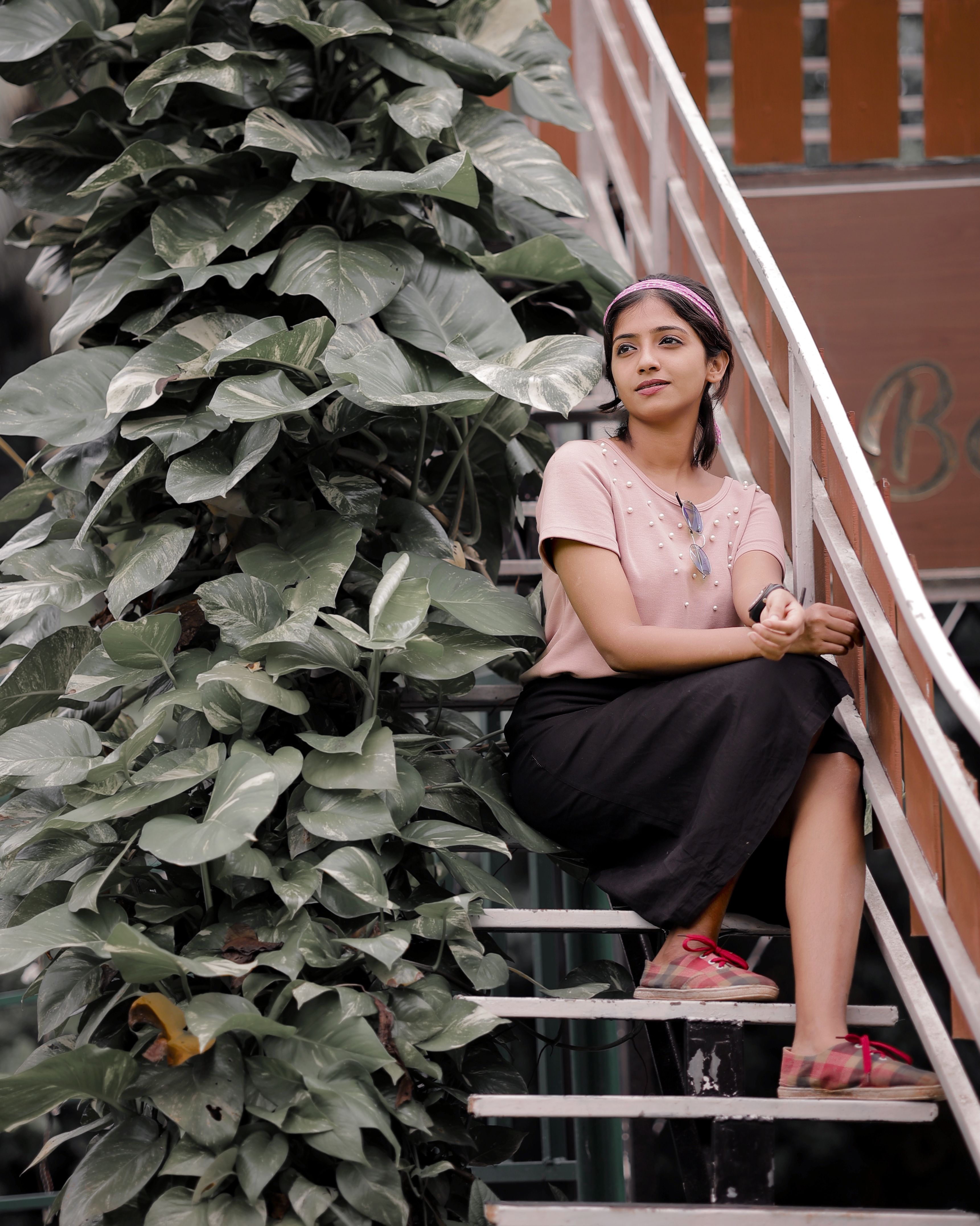 A young woman sits calmly on outdoor stairs next to lush green leaves, enjoying a peaceful moment.