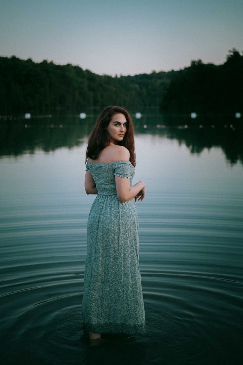 Free A woman in a dress stands in a tranquil lake at dusk, exuding calm beauty. Stock Photo