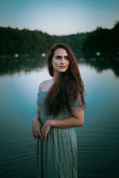 Free A serene portrait of a woman in a dress standing by a tranquil lake at dusk, exuding elegance. Stock Photo