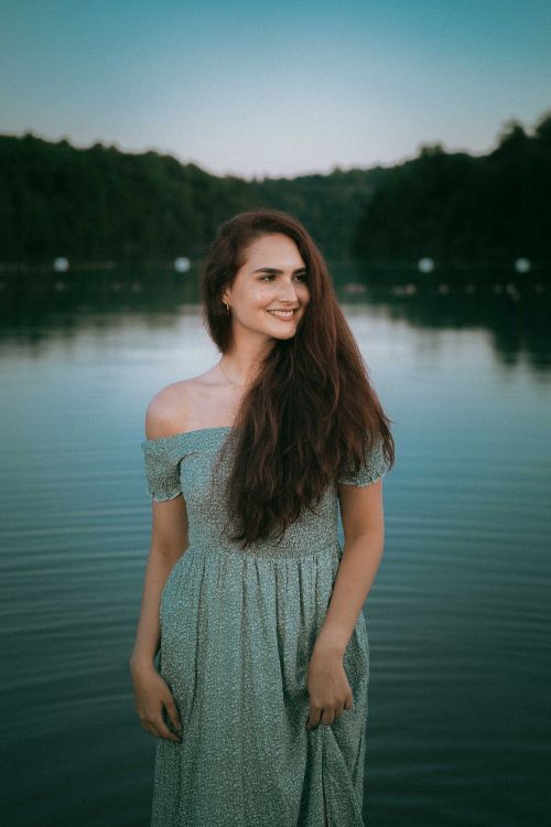 Free A brunette woman smiles standing by a serene lake in a flowing dress, captured at dusk. Stock Photo