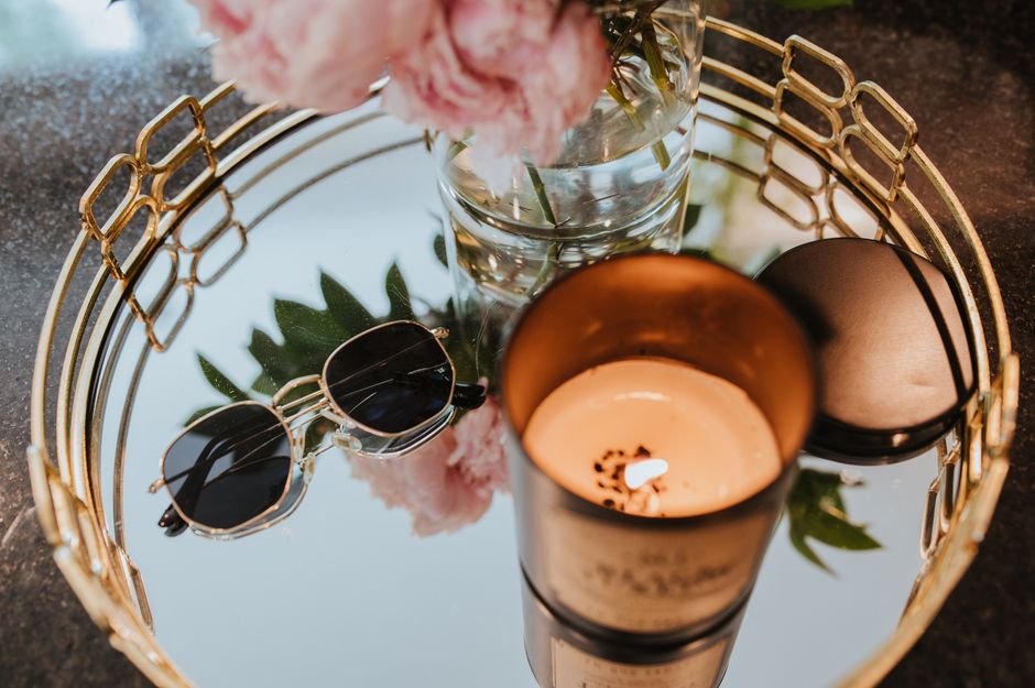 Mirror tray with a lit candle, sunglasses, and flowers in a vase creating a cozy atmosphere.