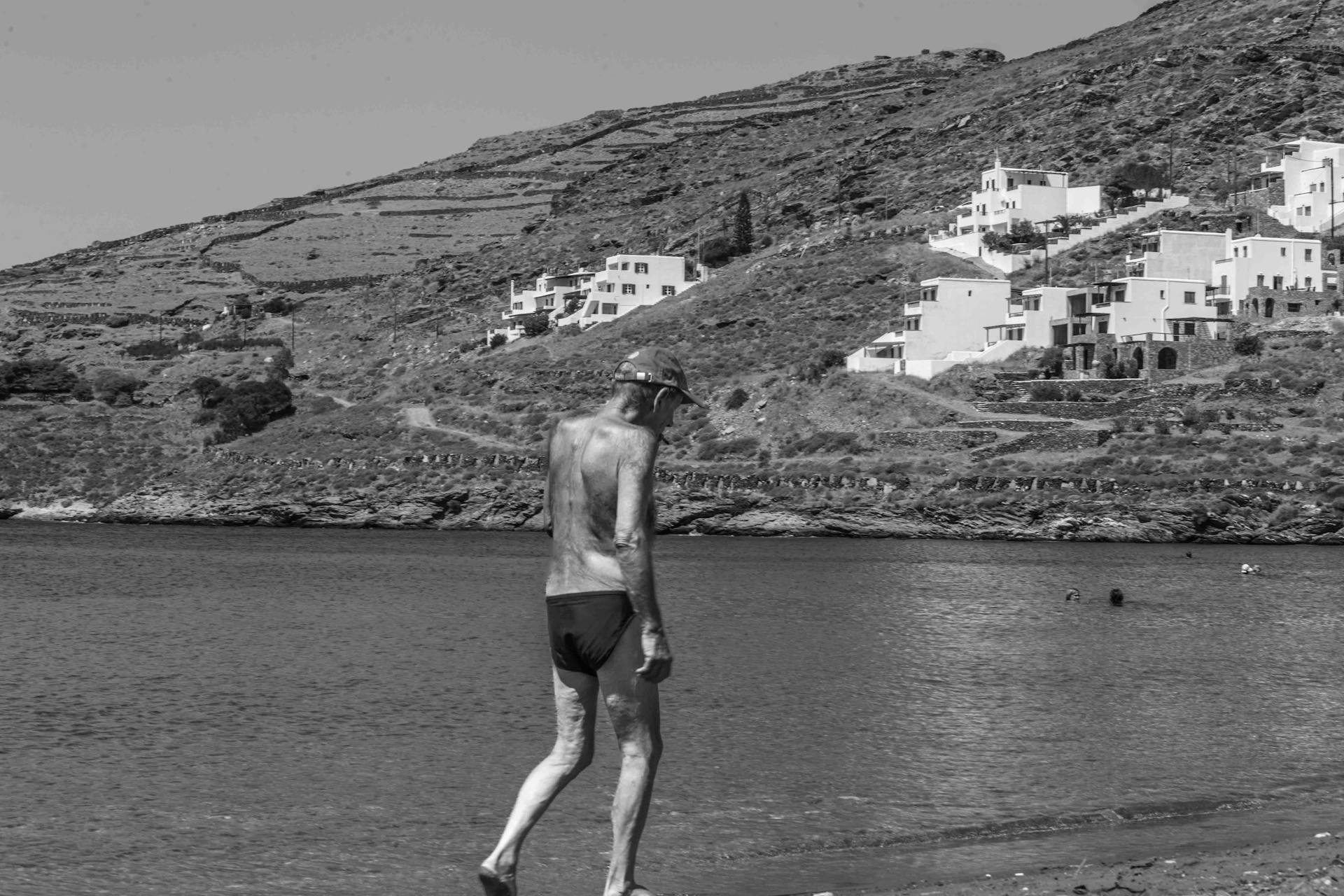 A senior man strolls along a tranquil beach, set against a hillside village backdrop in black and white.