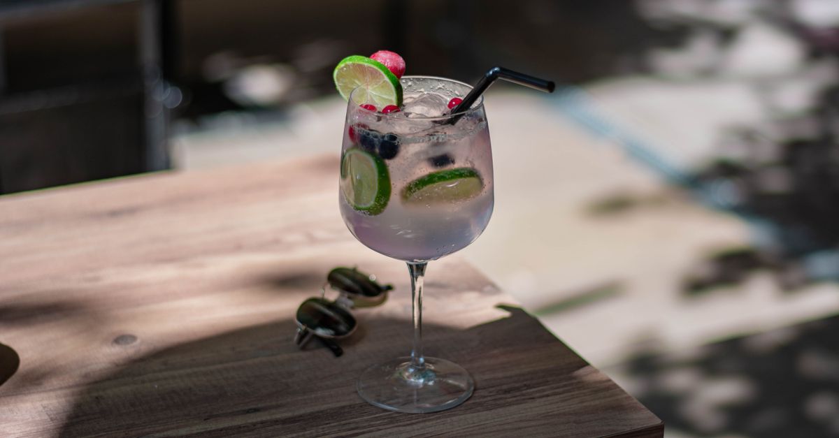 A refreshing gin cocktail garnished with lime and berries on a wooden table outdoors in sunlight.