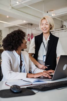 Two professional women engaging in teamwork using a laptop in a bright, modern office space.