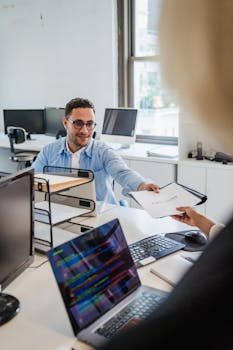 Two office workers exchanging documents in a bright office setting.