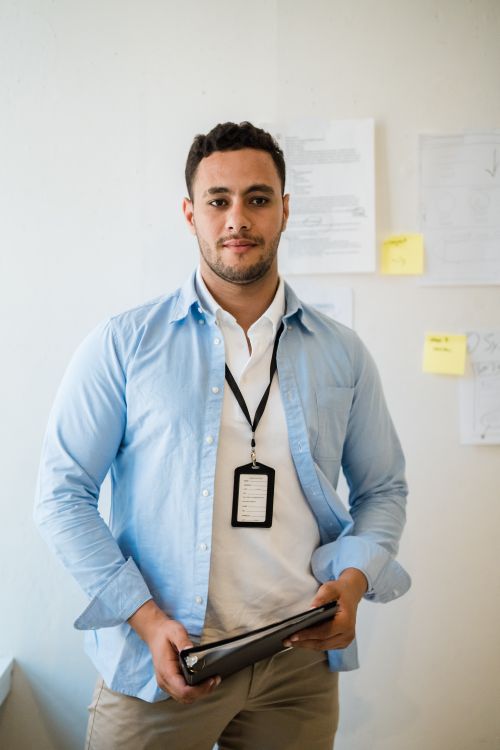 Free Portrait of a confident young male employee holding a notebook in an office environment with documents on the wall. Stock Photo