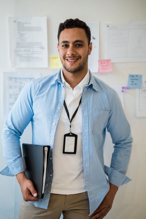 Free Smiling man in a blue shirt holding a binder, posing in an office environment. Stock Photo
