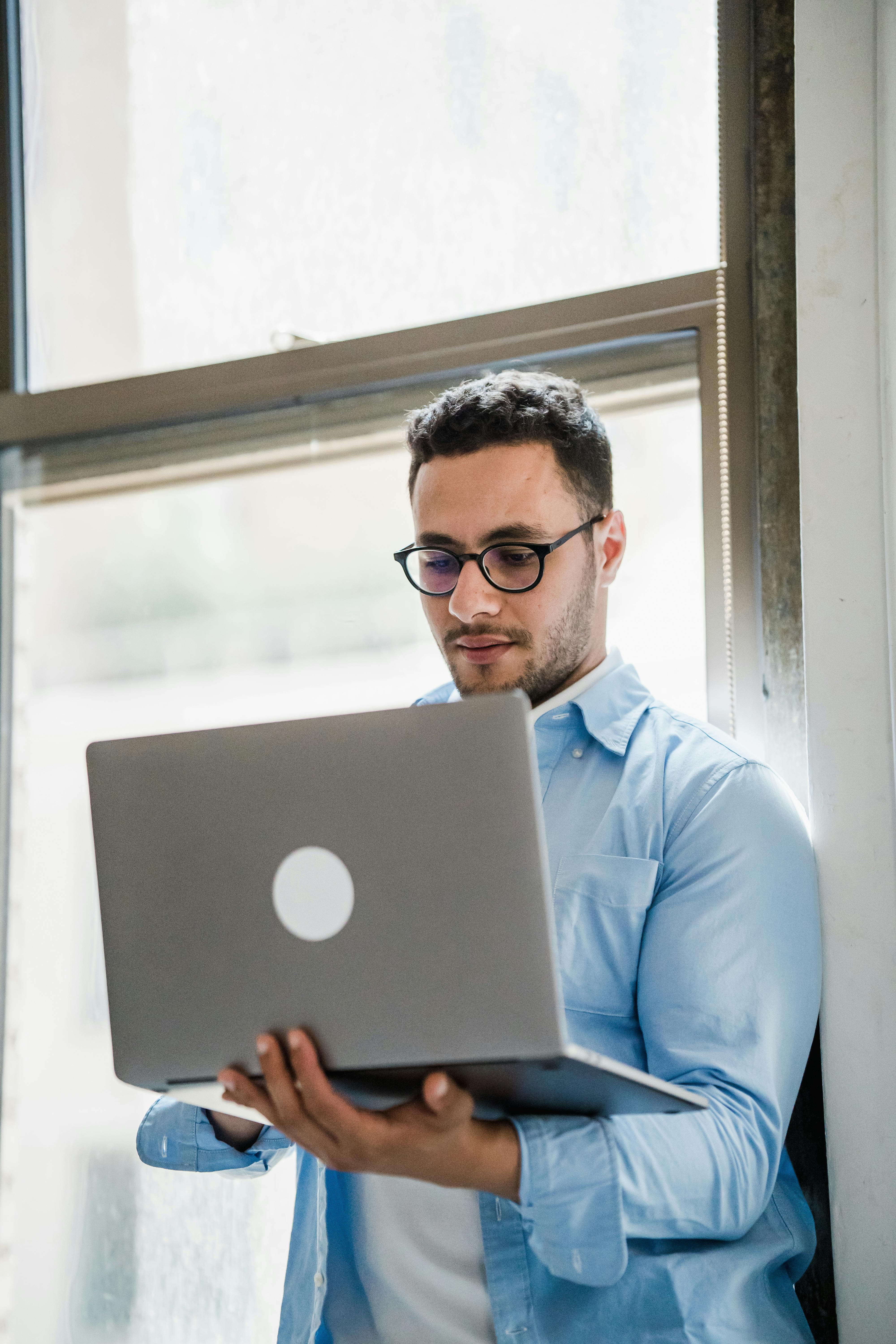 Office Worker Standing by the Window in an Office and Using his Laptop · Free Stock Photo