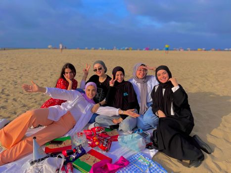 Group of women having a relaxed picnic on the sandy beach of Martil in Morocco, wearing traditional and casual attire.