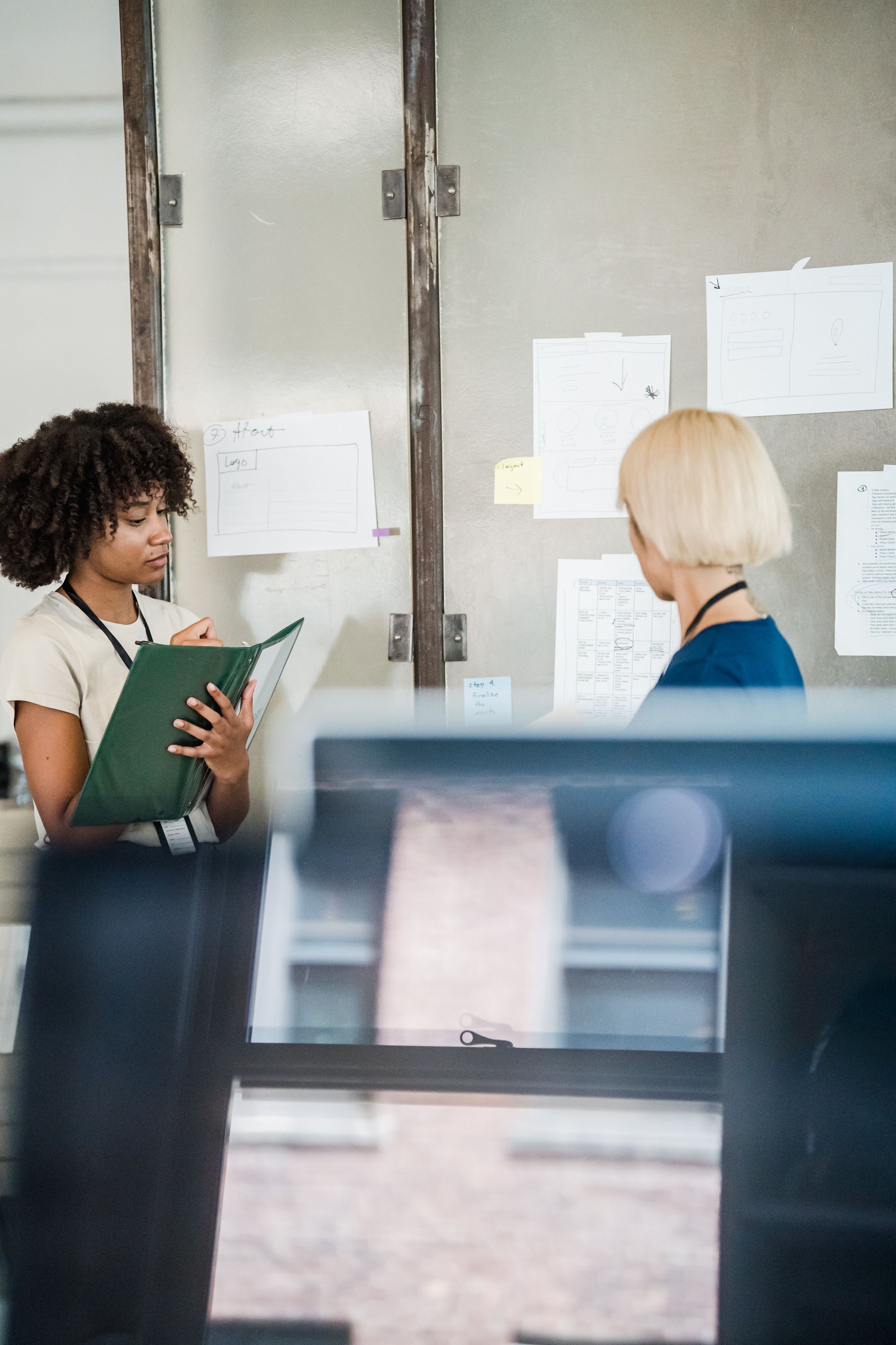 Two colleagues discussing a process in an office, with working documents pinned to the wall