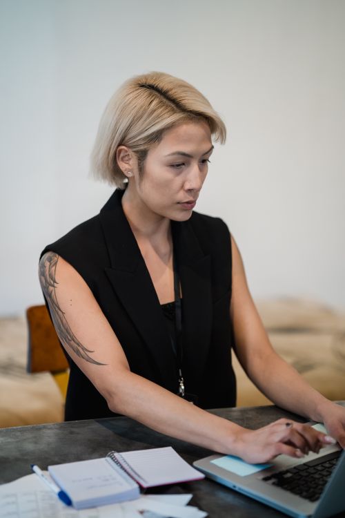Free Blonde woman with tattoo working on laptop in office setting. Stock Photo