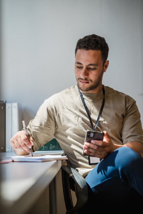 Free Man Making Notes Using Cellphone at Workplace Stock Photo